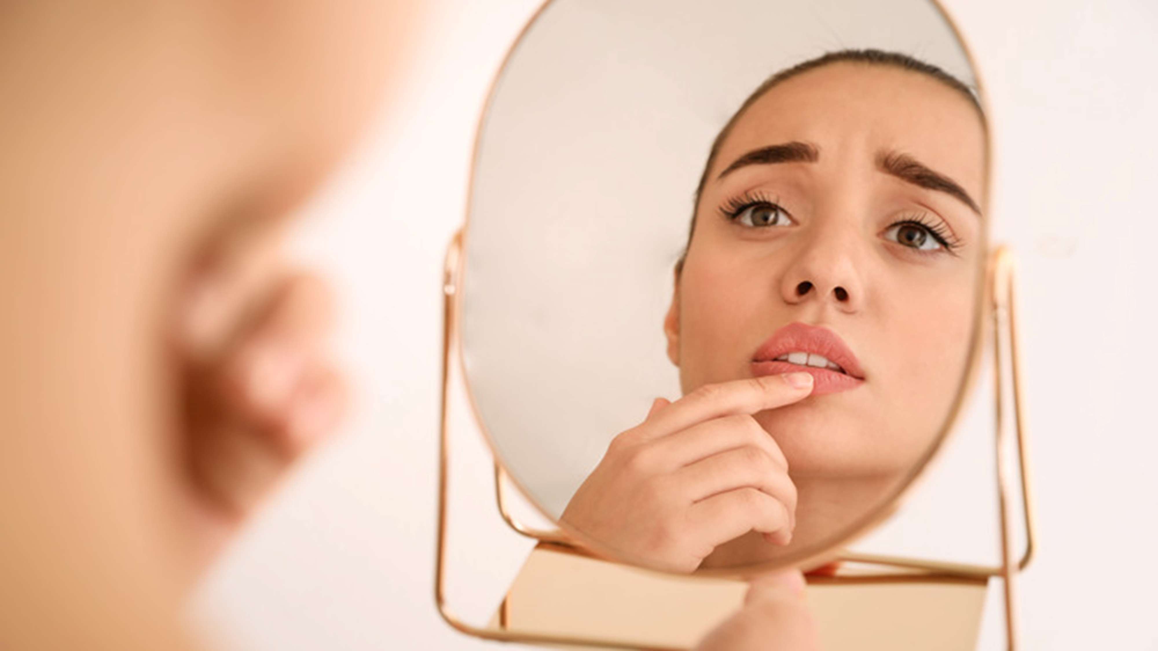 A woman looks at her reflection in a handheld mirror, touching her lips with concern as she examines a possible cold sore.